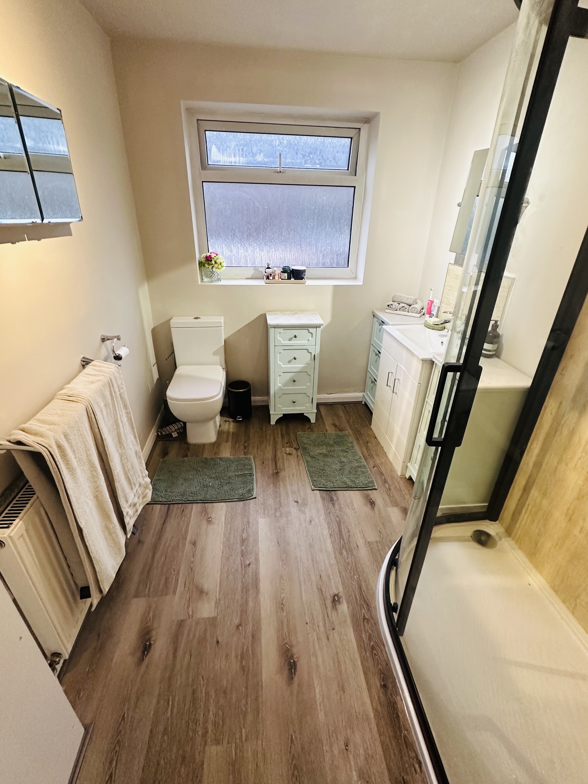 A bathroom with wood effect flooring, a cream drawer unit and a black framed shower screen